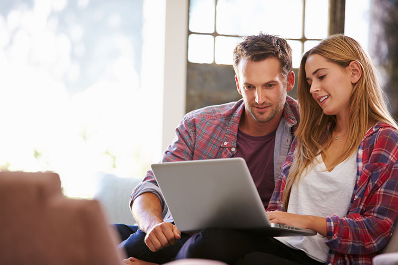 A man and a woman sit together on a couch, smiling and looking at a laptop screen. They appear relaxed and engaged, with natural light coming through a window behind them.